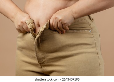 Middle-aged woman having large abdomen putting jeans on with hands on beige background. Visceral fat. Body positive. Sudden weight gain. Tight little clothes. Need for wardrobe change.
