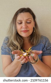 Middle-aged Woman Holding Gold And Silver Star Shapped Birthday Confetti In Hands While Standing Against Beige Wall, Mature Female Preparing Decorations For Special Event. Party Decor Concept