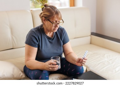 Middle-aged woman holds a blister of pills and reads medical instructions sitting on the couch - Powered by Shutterstock