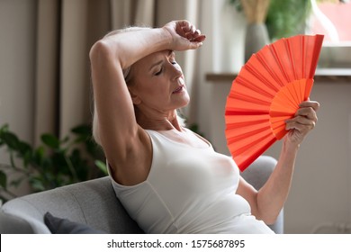 Middle-aged Woman Leaned On Sofa Resting In Living Room Cools Herself With Orange Colour Hand Fan Touch Forehead Feels Unwell Suffers From Unbearable Hot Summer Weather At Home Without Airconditioner