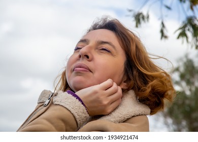 Middle-aged Woman In A Light Sheepskin Coat Stretches Her Face Towards The Sky With Her Eyes Closed Outside