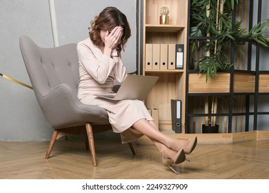 Middle-aged woman in a long beige dress sits on an armchair with a laptop on her lap. The woman covered her face with her hands in despair. Problems of the head, mistakes in business. - Shutterstock ID 2249327909