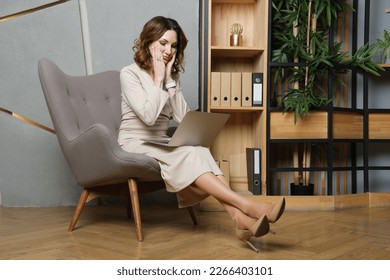 Middle-aged woman in a long beige dress sits on an armchair with a laptop on her lap. The woman covered her face with her hands in despair. Problems of the head, mistakes in business. - Shutterstock ID 2266403101