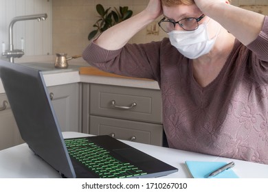 A Middle-aged Woman In A Medical Mask And Glasses For A Laptop In Cozy Kitchen Expresses Emotions Of Panic And Anxiety. Self-isolation, Remote Work, Anxiety. Horizontal Orientation, Selective Focus