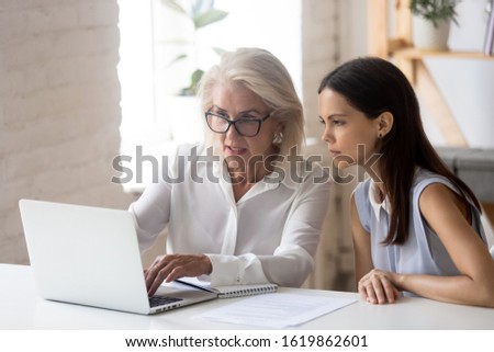 Middle-aged woman mentor explain to apprentice intern new corporate application seated at desk looking at computer screen communicating solving issues, planning project, mentoring and coaching concept