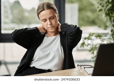 Middle-aged woman with neck pain, stressed, sitting in front of laptop.