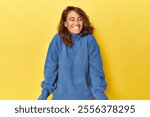 Middle-aged woman on a yellow backdrop laughs and closes eyes, feels relaxed and happy.