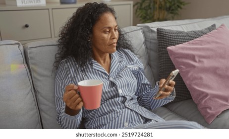 Middle-aged woman in pajamas holding a cup and looking at her phone while sitting on a cozy couch in a living room, with a relaxed and thoughtful expression. - Powered by Shutterstock