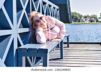 Middle-aged Woman In A Pink Hoodie Resting On A Pantone Bench At The Lakeside, Smiling And Looking At The Camera