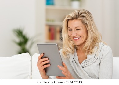 Middle-aged Woman Reading A Message, E-book Or Information On Her Tablet Computer With A Look Of Excited Anticipation As She Sits On A Couch At Home