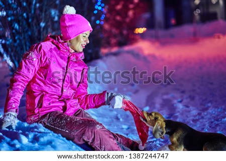 A middle-aged woman in a red sportswear is having fun playing with a dog puppy on a winter holiday Christmas night.