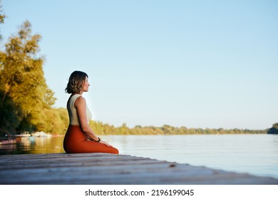 A Middle-aged Woman Is Relaxing On The Dock And Doing Yoga Breathing Exercises. She Put Her Feet In The Water.