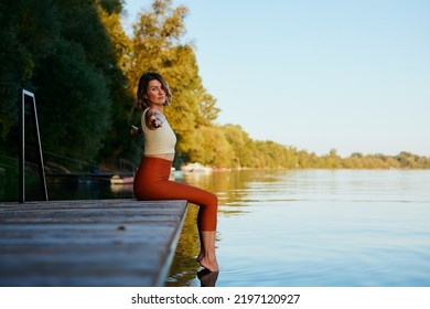 A Middle-aged Woman Is Relaxing On The Dock And Doing Yoga Breathing Exercises. She Put Her Feet In The Water.