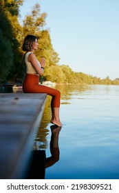 A Middle-aged Woman Is Relaxing On The Dock And Doing Yoga Breathing Exercises. She Put Her Feet In The Water.