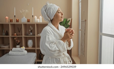 Middle-aged woman relaxing in a spa room, wearing a bathrobe and towel on her head, holding a cup, with candles and plants in the background - Powered by Shutterstock