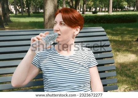 Middle-aged woman with a short haircut drinks water from a plastic bottle in the city park. The need to drink fluids while in the heat.
