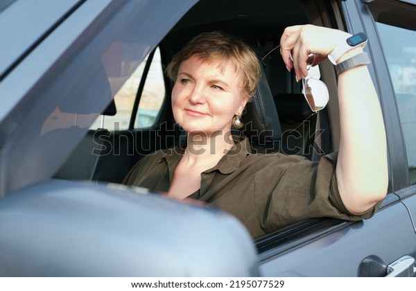 Middle-aged woman with a short haircut\
sits in her car behind the wheel and looks away dreamily. Woman\
holds sunglasses in her hands. Woman is driving a\
car.