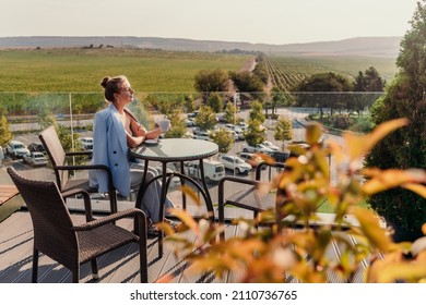 A middle-aged woman sits in a street cafe overlooking the mountains at sunset. She is dressed in a blue jacket and drinks coffee admiring the nature. Travel and vacation concept.