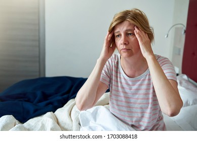 A middle-aged woman is sitting on the bed, holding her head. Health problems, headache, menopause, insomnia.
