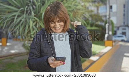 A middle-aged woman smiles while using a smartphone outdoors in casual attire, exuding joy and modern connectivity.