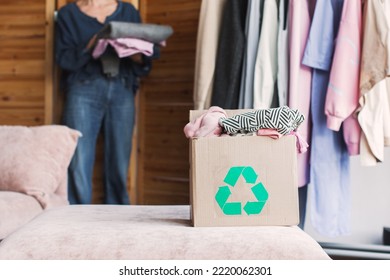 Middle-aged Woman Sorting Old Clothes Into Box With Recycle Sign. Green Recycling Sign On Box. Woman Throwing Out Old Clothes.