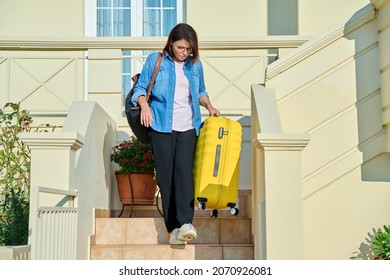 Middle-aged woman with suitcase and backpack walking down steps of house