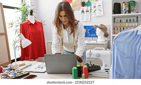 A middle-aged woman tailoring in a design room, focused on using her laptop surrounded by colorful threads and fabrics. - Powered by Shutterstock