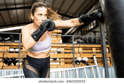 A middle-aged woman is training with a punching bag. The 50-year-old is hitting with her left arm with a boxing glove. Concept of female boxers between 40 and 50 years old. Boxing for adult women. - Powered by Shutterstock