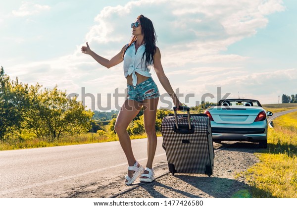 Middle-aged\
woman with a travel bag hitchhiking on the narrow empty rural road\
in summer on the background of a broken car beautiful nature\
outside the city. car problems on the\
road