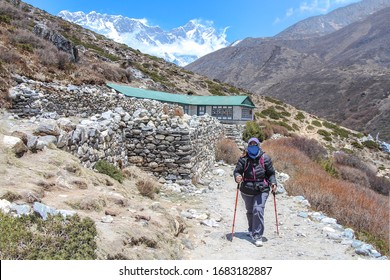 Middle-aged Woman Walks On Footpath From Everest Base Camp Next To Typical Guest House In Himalayas. Everest And Lhotse Mountain Peaks Are Visible In The Background. Trekking In Nepal Theme.