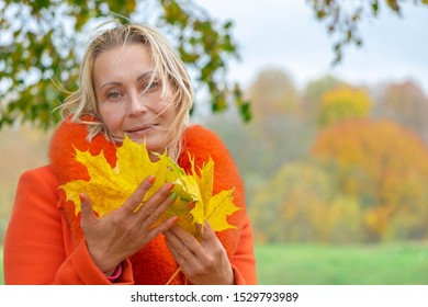 Middle-aged Woman With Wind Waving Hair In Orange Coat Holding Autumn Maple Leaves In Colorful Fall Nature Background. Portrait Of Romantic Female