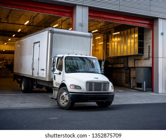 The middle-duty white day cab compact semi truck for local shipping and delivery services with long box trailer leaves the gate of multistory building after unloading the delivered cargo.