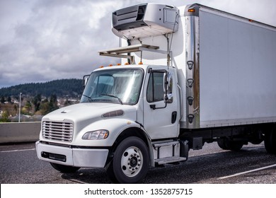 The Middle-duty White Day Cab Compact Semi Truck For Local Shipping And Delivery Services With Refrigerated Box Trailer Running On The Wide Highway To Deliver Loaded Chilled Fresh Produce