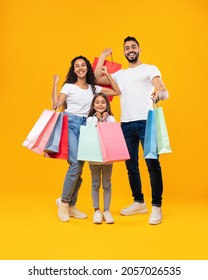 Middle-Eastern Family Holding Shopping Bags Smiling To Camera Standing On Yellow Studio Background. Parents And Daughter Posing With Colorful Paper Shoppers. Sales Season Advertisement. Vertical