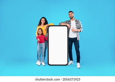 Middle-Eastern Family Standing Near Full-Length Phone With Blank Screen Gesturing Thumbs Up Posing On Blue Background. Arabic Parents And Daughter Leaning At Cellphone Approving Great Application