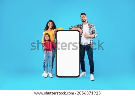 Middle-Eastern Family Standing Near Full-Length Phone With Blank Screen Gesturing Thumbs Up Posing On Blue Background. Arabic Parents And Daughter Leaning At Cellphone Approving Great Application