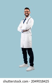 Middle-Eastern Male Doctor Posing Wearing White Uniform Crossing Hands Over Blue Background, Smiling To Camera. Full Length Shot Of Professional Medical Worker, Successful Physician