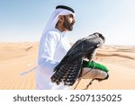 Middle-eastern man wearing traditional emirati arab kandura in the desert and holding a falcon bird - Arabian muslim adult person at the sand dunes in Dubai