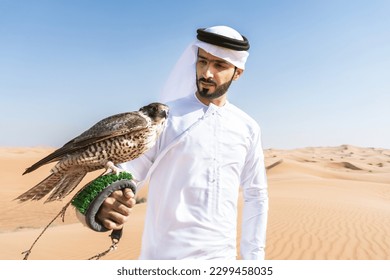 Middle-eastern man wearing traditional emirati arab kandura in the desert and holding a falcon bird - Arabian muslim adult person at the sand dunes in Dubai - Powered by Shutterstock