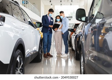 Middle-eastern Young Man And Woman In Protective Face Masks Choosing Auto In Dealership Salon While COVID-19 Pandemic, Having Conversation With Sales Manager, Reading Car Specifications, Full Length