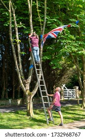 Middleton On Sea, Sussex / UK - 7 May 2020: An Older Man Climbs A Tall Ladder 18 Feet (6 Metres) Without Safety Gear To Hang Red, White And Blue Bunting And Flags In A Tree. His Friend Gives Support.