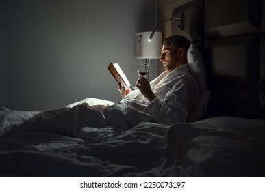 Midle-aged man relaxing rin bed reading book holding a glass of red wine with bedside lamp turned on. Evening relaxation, hobbies, free time concept. Adulthood concept.