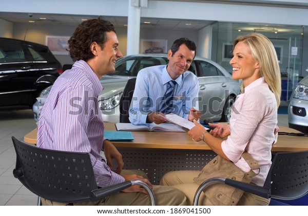 Mid-length shot of a car salesman and couple\
sitting at the desk in a large car showroom with the woman is\
handing over\
paperwork.