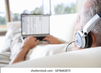 Mid-length shot of a senior man relaxing on the sofa at home listening to music on headphones using the laptop.