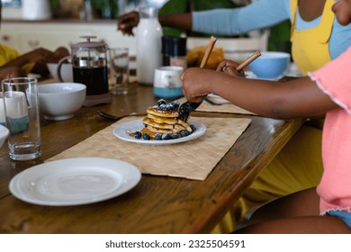 Midsection of african american girl eating pancakes with fork and table knife at dining table. Unaltered, lifestyle, childhood, morning, breakfast, blueberry, hand and home concept. - Powered by Shutterstock