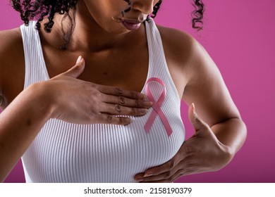 Midsection Of African American Mid Adult Woman With Pink Breast Cancer Awareness Ribbon On Breast. Pink Background, Breast, Cancer, Medical, Ribbon, Awareness, Support, Healthcare And Alertness.