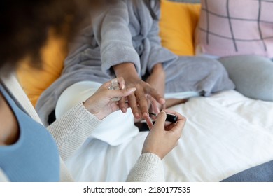 Midsection Of Biracial Young Woman Painting Female Friend's Fingernails While Sitting On Bed At Home. Body Care, Pampering, Unaltered, Friendship, Togetherness And Lifestyle Concept.