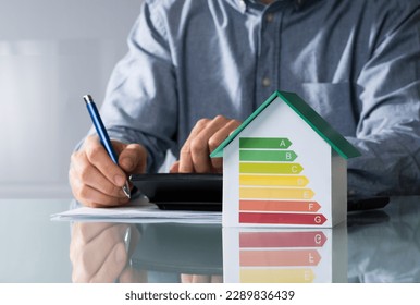 Midsection Of Businessman Calculating Invoice With Energy Efficient Chart On House Model In Office