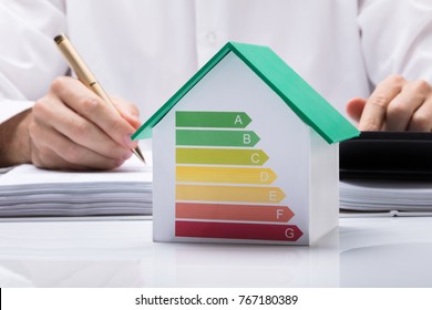 Midsection Of Businessman Calculating Invoice With Energy Efficient Chart On House Model In Office