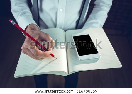 Midsection of businessman using smartphone while writing in his notebad at office